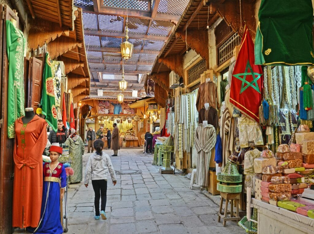 Explore the bustling and colorful market street in Fès Medina, filled with traditional Moroccan goods and attire.