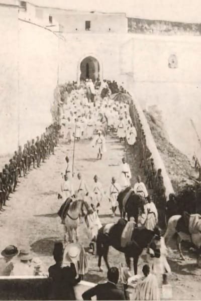 A historical photograph (late 19th century) showing a large crowd of locals in traditional attire gathered at a grand entrance to the Fez medina (representing Bab Bou Jeloud). The scene captures the bustling hub status of the historic gateway.