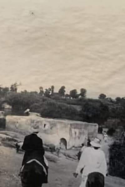 A large, imposing historical gate (Grand Entrance) to the Fez Medina, featuring a sturdy stone arch and ancient architecture. The scene evokes the passage of merchants and scholars from the late 19th century.