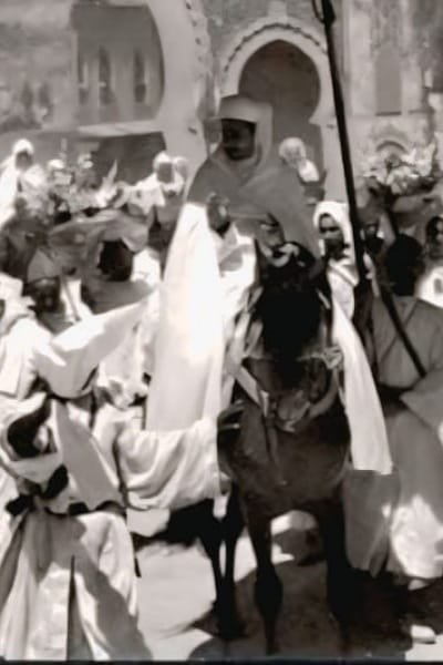 A historical image of King Mohammed V in Fez, riding a horse in a procession and surrounded by crowds. The scene symbolizes his strong leadership and the deep connection between the monarch and the Moroccan people.