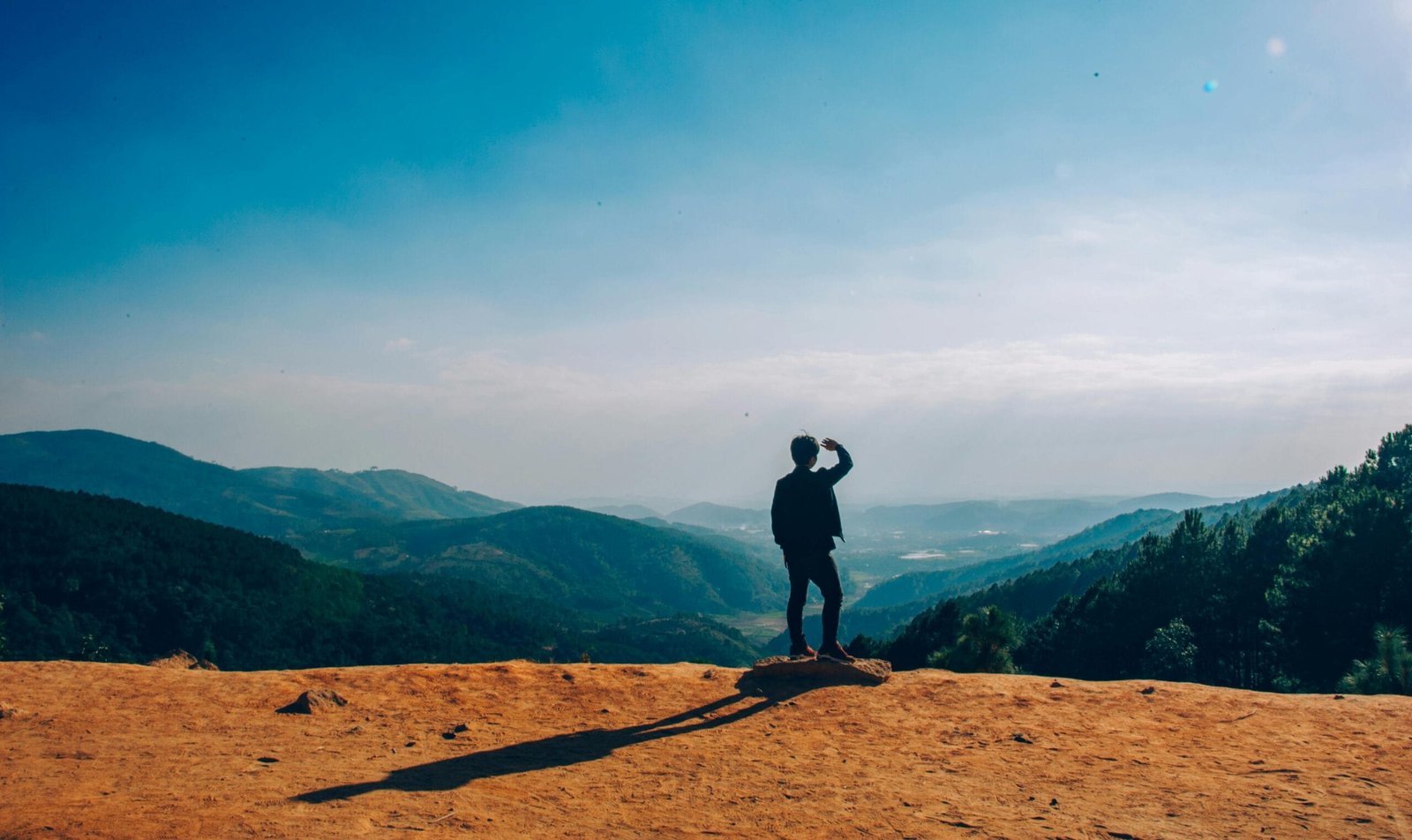 Custom Transfers 2 A lone traveler stands on a hilltop, gazing over a vast mountain landscape under a clear blue sky.
