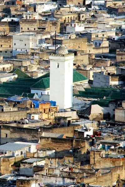 A vibrant, narrow souk alley in the Fez medina, showcasing traditional wooden coverings and architectural elements. The scene highlights the unique juxtaposition of the old-world charm (architecture) with the bustling urban life (chaos) in Fez's marketplace.