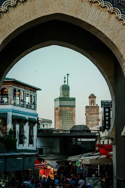 A photograph taken through the ornate arch of Bab Boujloud (The Blue Gate), framing the famous minarets of the old medina against the sky. The scene captures the essence of Fez, showcasing the iconic architectural entrance and the bustling life within the historical walls.