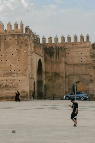 A panoramic view of Fez El Bali at sunset, showcasing the medieval walls in the foreground and the densely packed ancient medina in the distance. The scene is painted in rich, golden hour tones, evoking history and nostalgia.