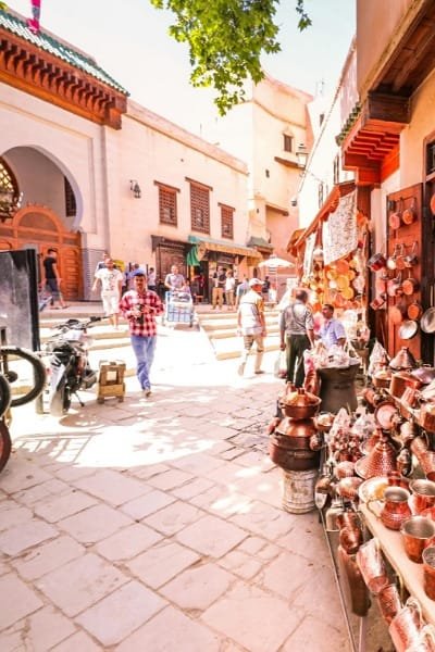 A bustling scene at Place Seffarine in Fez, showing a coppersmith selling wares and traditional work in the square named after the 'saffarine' artisans. The image captures the living heritage of craftsmanship in the heart of Fez's old medina.