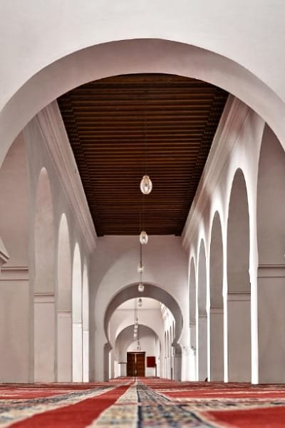 A peaceful scene in the courtyard (sahn) of a historic Moroccan mosque, possibly the Andalusian Mosque in Fez, featuring a central ablution fountain surrounded by traditional decorative arches and stucco work. The atmosphere provides a historically rich experience.