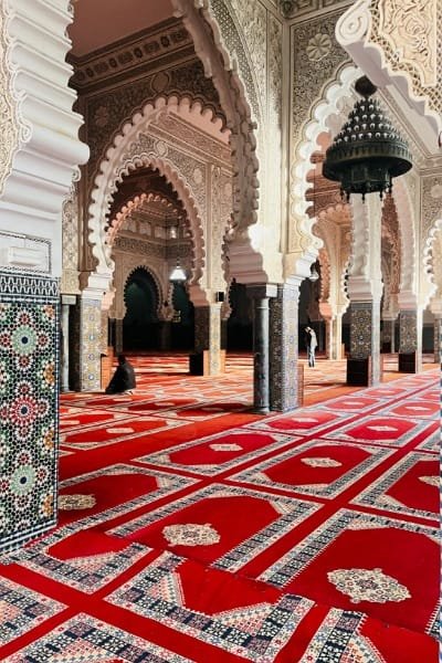 Interior view of a Moroccan mosque, potentially the Andalusian Mosque in Fez, featuring intricate stucco work, ornate arches, and geometric Zellige tiles. The architecture embodies the unique Andalusian-Moroccan heritage.