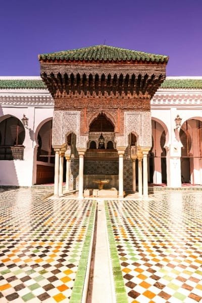 The ornate central courtyard (Sahn) of the Mosque of the Andalusians in Fez, featuring a richly decorated fountain pavilion, detailed zellige tilework, and surrounding white horseshoe arches. This architecture reflects the deep Andalusian-Moroccan cultural heritage of the 9th-century mosque.