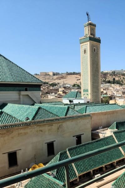 A prominent view of the decorated minaret of the Mosque of the Andalusians in Fez, rising above the surrounding traditional green-tiled roofs of the old medina. This landmark, founded in 859 CE, showcases the beautiful Andalusian-Moroccan architectural fusion.