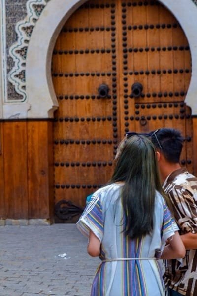 An imposing, traditional Moroccan wooden door (likely belonging to the Nejjarine Fountain or Fondouk Nejjarine), intricately decorated with brass studs, framed by a stucco arch in Nejjarine Square, the historical heart of carpentry craft in Fez.