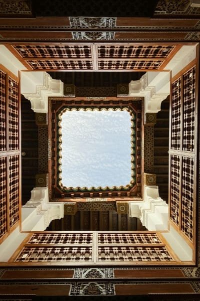 A beautiful, traditional Moroccan wooden door with a horseshoe arch and brass studs, framed by intricate stucco and colorful zellige tiles, representing the carpentry artistry of Nejjarine Square in Fez.