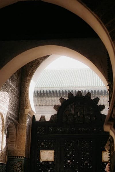 A stunning view through a traditional Moroccan archway inside the Bou Inania Madrasa, highlighting an intricately carved wooden door and ceiling details. The architecture reflects the grandeur of the 14th-century Marinid design.