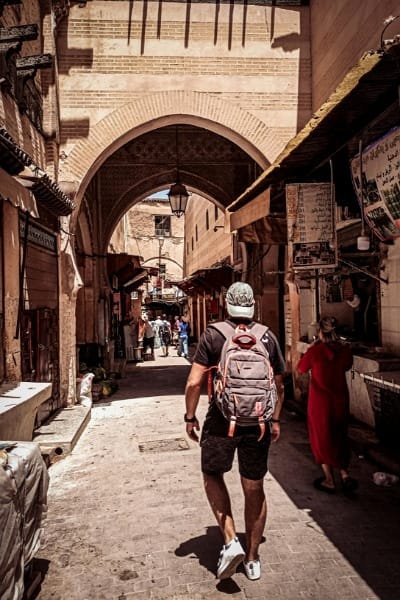 A low-angle shot of a female traveler looking up at the intricate 14th-century Moroccan artistry inside the Bou Inania Madrasa, showcasing geometric zellige tiles and elaborate stucco work in the courtyard. The site requires a small entrance fee to view this historical art.