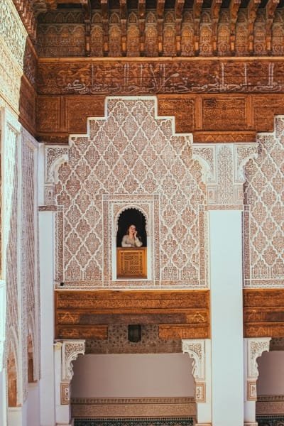 A stunning view inside the Attarine Madrasa in Fez, showing a woman in an upper-level window surrounded by richly carved cedar wood and delicate geometric stucco patterns. This 14th-century religious school is a masterpiece of Moorish architecture.
