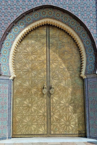 The massive, ornate golden brass gate of the Royal Palace (Bab Makina) in Fez, featuring intricate geometric star patterns and framed by a colorful zellige arch. The palace symbolizes Morocco’s royal splendor and is near the historic Jewish Mellah.