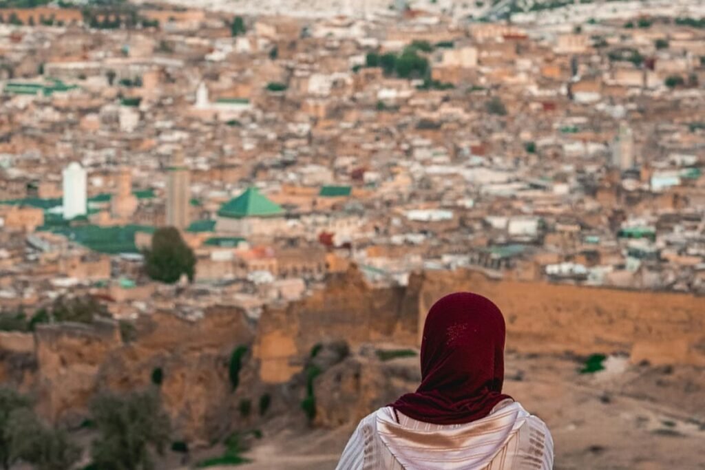 A panoramic view of the vast Fez Medina from a hilltop, showing crowded rooftops, ancient alleyways, and the green-tiled roofs of mosques. This image showcases the immense scale and UNESCO-listed beauty of the old city.