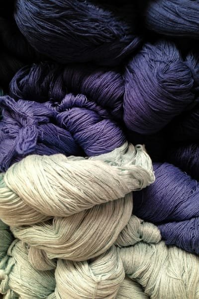 A close-up of piles of rich, naturally dyed Moroccan yarns, featuring vibrant shades of deep indigo and soft purples and grays. This wool is representative of the hand-dyed textiles found in the Souk as-Sabbaghine (Dyeing Quarter) in Fez.