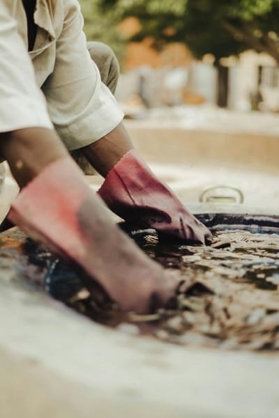 A close-up of a craftsman's hands wearing gloves, dipping materials into a traditional dyeing vat at Souk as-Sabbaghine in Fez. The image highlights the ancient, manual techniques used to apply natural dyes to textiles and leather.
