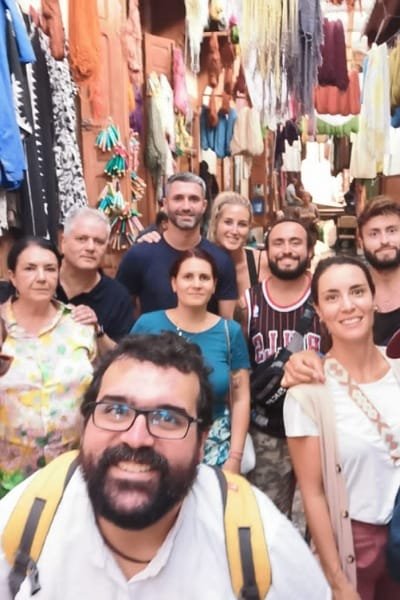 A group of tourists smiling in the colorful Souk as-Sabbaghine (Dyeing Quarter) in Fez, surrounded by vibrant hand-dyed Moroccan textiles hanging overhead. The image captures the lively, ancient craftsmanship using natural dyes.