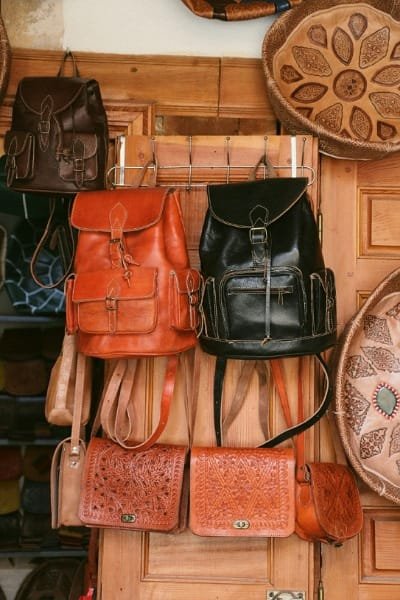 A display of authentic Moroccan leather goods, including various handmade backpacks and carved leather shoulder bags. These items are made from high-quality, durable leather processed at a Fez tannery like Chouara.