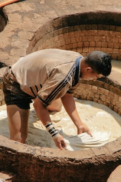 A leather tanner in Fez bending over a traditional vat, using manual methods passed down through centuries to prepare and dye leather. The scene captures the dedication to ancient craftsmanship and physical labor.