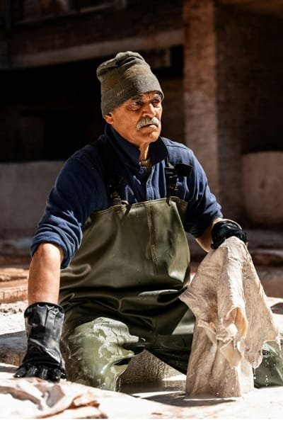 A leather craftsman at the Chouara Tannery in Fez preparing a raw hide in the initial stages of tanning, likely using a lime solution. The tanner wears a traditional straw hat, emphasizing the manual labor and ancient methods before the leather is dyed.