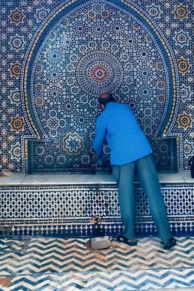A craftsman tending to a traditional Moroccan wall fountain in Fez, beautifully adorned with intricate blue and yellow Zellige tiles. The hand-cut geometric patterns showcase the highest quality of this centuries-old artistry.