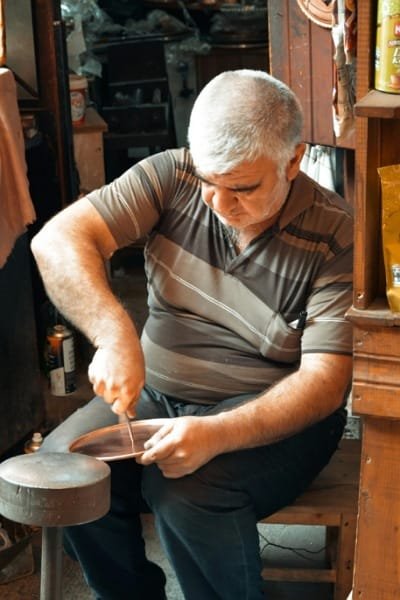 A skilled Moroccan artisan in Fez meticulously hand-engraving intricate arabesque patterns onto a copper tray. This ancient craft requires incredible skill and precision and is prominent near Njarine Square.