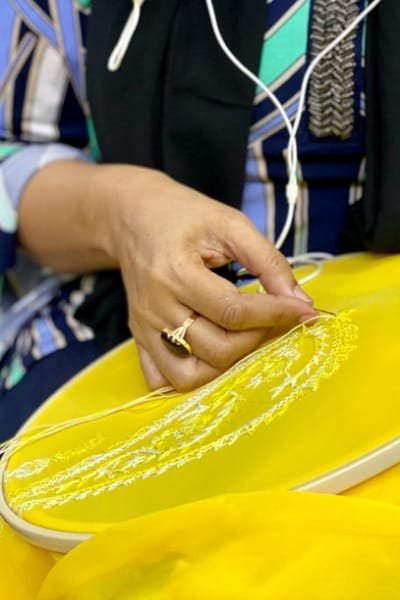A close-up of a skilled artisan's hands meticulously working on Fez embroidery, hand-stitching intricate designs onto bright yellow fabric with silk threads. The image highlights the dedication, precision, and skill required for this highly refined traditional Moroccan craft.