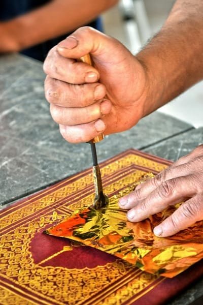 A close-up of a skilled leather artisan in Fez using a traditional tool for hand-engraving or gold stamping a complex pattern onto a piece of red leather. This process showcases the meticulous care and individual touch applied to creating unique, durable Moroccan leather accessories.