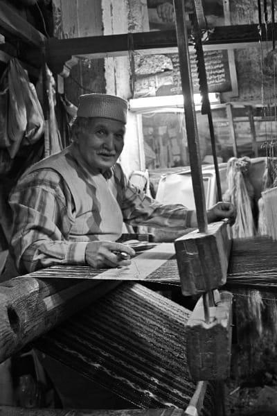 A smiling, traditional Moroccan weaver artisan in Fez working diligently on a large, traditional wooden loom. The image highlights the inherited skill and patience required for weaving Moroccan textiles in the old medina.