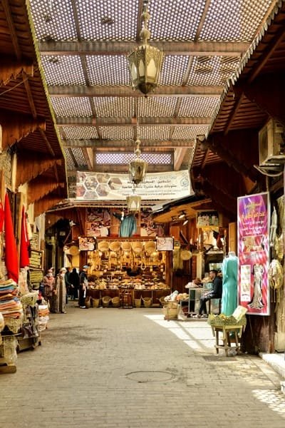 A wide, covered main road in the Fez Medina souk, filled with shops and showing high pedestrian traffic. The scene captures the welcoming and easily navigable arteries of the city that connect key historical landmarks.