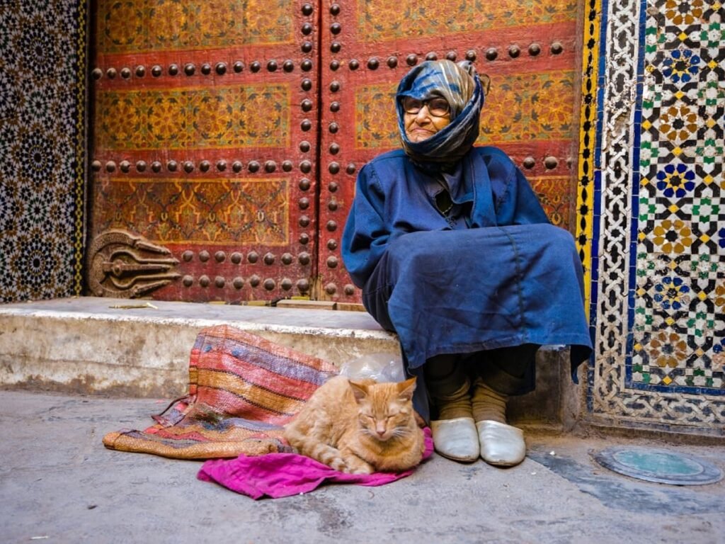 An authentic scene capturing the secrets of Fez, showing an elderly woman sitting quietly in front of an ornately decorated historical door. The image highlights the unique culture, detailed architecture, and serene hidden corners of the ancient city.