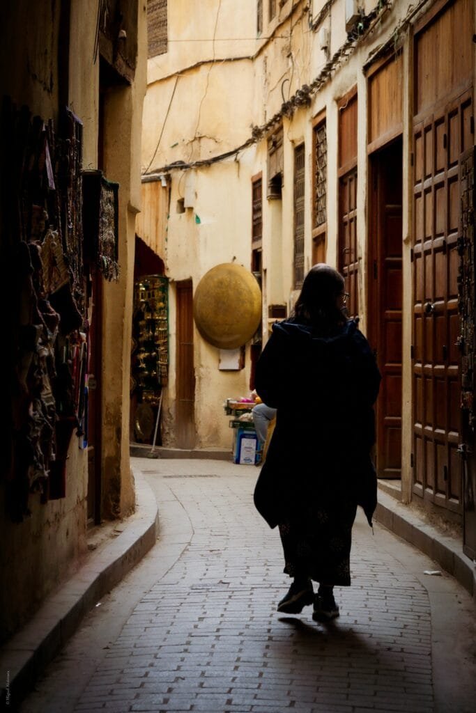 A local woman dressed in traditional attire walks through the narrow, maze-like streets of Fez — a living portrait of culture and everyday life.