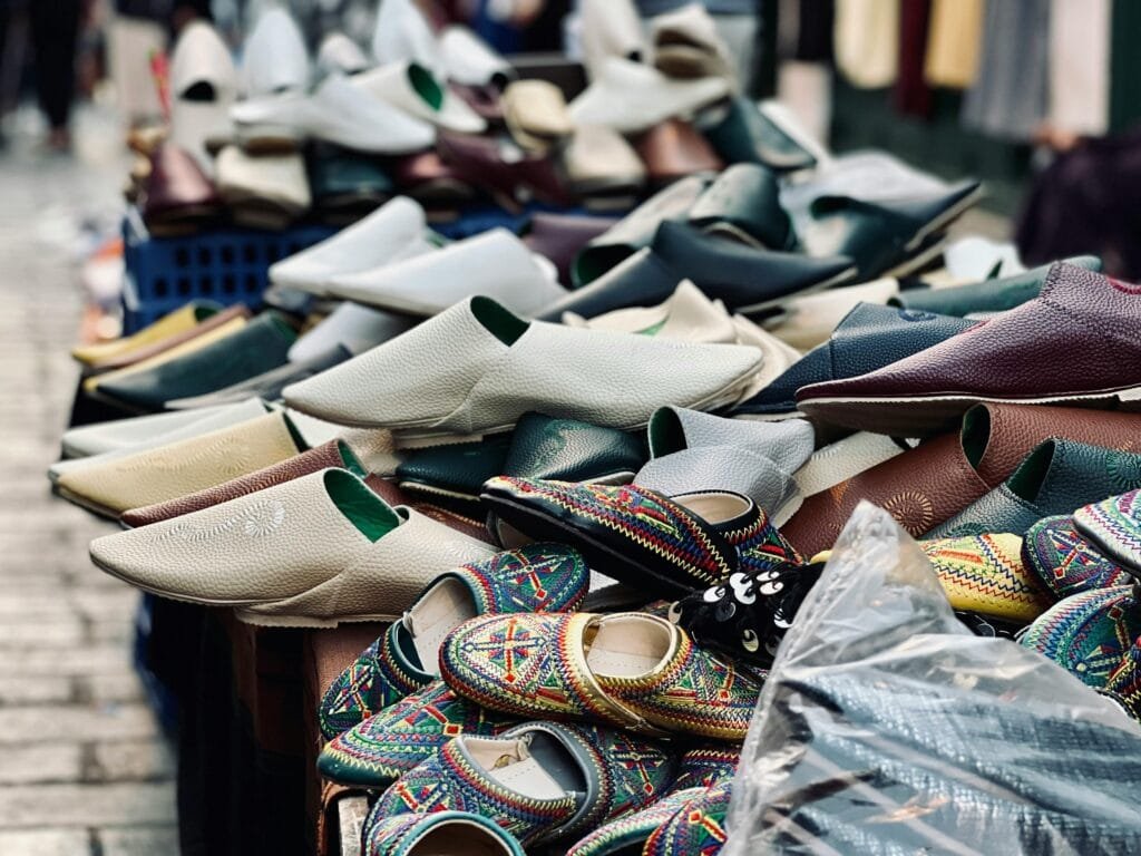 Fez Shopping Guide: What to Buy in Morocco’s Craft Capital 13 Ahlan wa sahlan to the captivating world of Fez, where ancient traditions come alive with every step! As you wander through the labyrinthine alleys of our magnificent medina, especially when you find yourself in the vibrant Qissariat Al-Kifah near the fragrant Attarine district, prepare to discover the soul of Moroccan craftsmanship embodied by the esteemed Kharrazine – our traditional leather shoemakers. More than just artisans, the Kharrazine are guardians of a time-honored legacy, their nimble hands transforming supple leather into exquisite footwear that graces the feet of Moroccans for every occasion. They are the creators of the iconic balgha, those elegant and comfortable leather slippers you see everywhere, and the beautifully adorned cherbil, the sophisticated footwear favored by Moroccan women. Imagine: each pair you see is a testament to centuries of skill, meticulously handcrafted without the aid of modern machinery. Feel the luxurious softness of the leather, often dyed with natural pigments that whisper tales of the earth. Admire the vibrant hues of the fabrics and the intricate stitching that adds a touch of artistry to every piece. But what truly makes these shoes extraordinary? A Touch of the Soul: Forget mass-produced uniformity. Each pair of Kharrazine creations possesses its own unique character, bearing the distinct mark of the artisan who poured their heart and expertise into its making. You are not just buying shoes; you are acquiring a piece of Moroccan artistry. Woven into Our Identity: These are not mere accessories; they are vibrant threads in the rich tapestry of Moroccan culture. From joyous celebrations and solemn weddings to the rhythm of daily life, these shoes are a tangible symbol of our heritage, our elegance, and our deep-rooted traditions. Wearing them is embracing a piece of Moroccan identity. Nature's Embrace: Only the finest, often naturally dyed leather is selected, ensuring unparalleled comfort and enduring quality. These are shoes designed not just to look beautiful, but to feel exquisite and last through time, becoming a cherished part of your own story. Your Journey to Discovery: To truly immerse yourself in this captivating craft, make your way to the Qissariat Al-Kifah (Victory Market). Here, within this traditional covered souk, a treasure trove awaits. Dozens of tiny stalls overflow with a dazzling array of handmade Moroccan slippers, from the elegantly simple to the lavishly ornate. A Tip for the Curious Traveler: Don't just let your eyes feast on the beauty before you – engage with the artisans! They are often delighted to share their craft, custom-fit a pair just for you, or even offer a glimpse into the intricate process of creation. Owning a pair of authentic Moroccan balgha is more than just acquiring a unique and stylish souvenir; it's forging a direct connection to the living heritage of Fez, a tangible reminder of the artistry and soul you encountered within its ancient walls. Walk away with a piece of our story, ready to create your own.