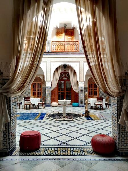 The inviting interior of a traditional Moroccan home salon (Sedari) featuring colorful Zellige mosaic tiles, and warm light filtering through intricate stained-glass windows. This scene captures the cozy and vibrant aesthetics of Moroccan decor.