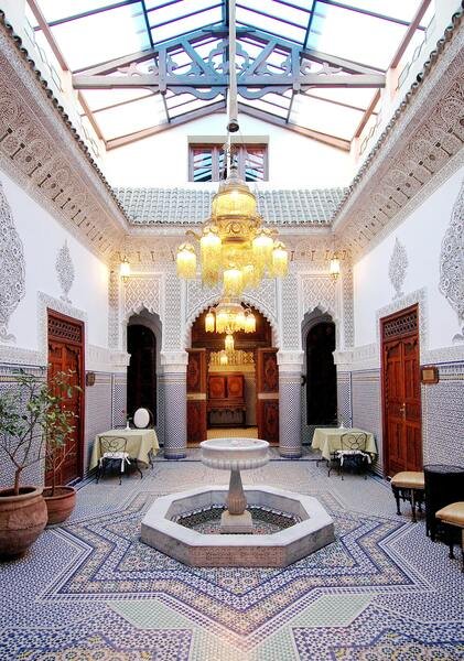 A stunning traditional Moroccan Riad courtyard featuring vibrant blue and white Zellige mosaic tiles, graceful arched doorways, and a central marble fountain. The image showcases the timeless beauty of Moroccan architecture.