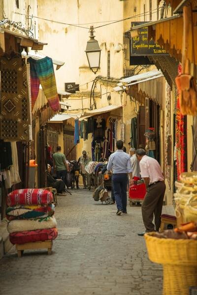 An open, narrow alley in the Fez Medina filled with pedestrians and goods, highlighting the connection to vibrant artisan workshops. This path is an excellent place to explore and get lost in the rhythm of daily life in Fez.