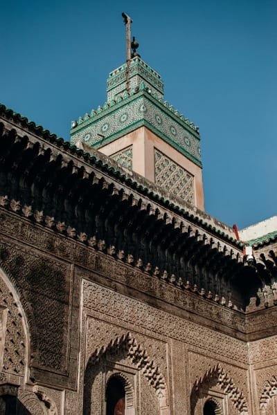 A detailed close-up of the minaret of the Bou Inania Madrasa in Fez, showcasing intricate green Zellige tiles and carved plasterwork. The madrasa, founded in the 14th century, is a prominent historical monument embodying traditional Islamic architecture and was a significant educational center.