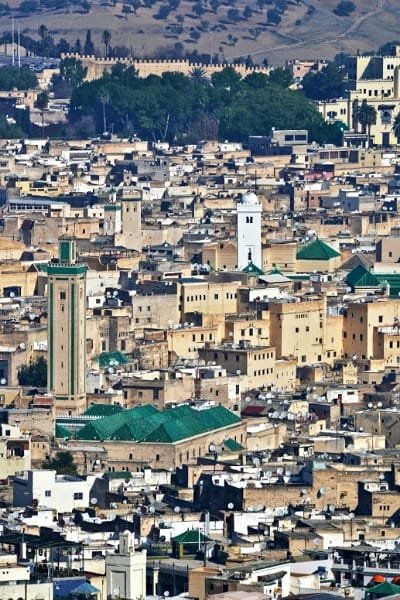 Tourists walk through an exquisitely decorated arched passageway in the Fez medina, featuring Zellige tiles and carved plasterwork. The image represents Fez as a city of living history and a treasure of Islamic architecture, contrasting with the fading traditional craftsmanship.