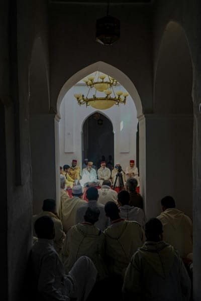 A group of men in traditional Moroccan attire sitting in a dimly lit, arched religious setting for a spiritual gathering or dhikr session. This image illustrates the profound religious atmosphere, Sufi rituals, and nightly spiritual traditions in the sacred city of Fez.