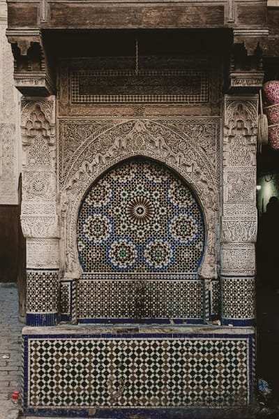 A detailed close-up of the Nejjarine Fountain in Fez, showcasing intricate Zellige tiles and carved stucco and wood in traditional Moroccan Islamic patterns. The fountain is a key highlight of Nejjarine Square, a historic center for woodworking and artisanal traditions.
