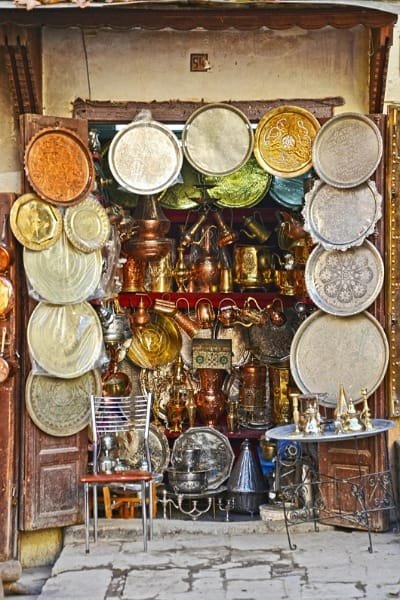 A brightly lit shop display in Fez showcasing a multitude of handcrafted Moroccan brass and copper items, including large trays and teapots. The products feature intricate geometric and floral designs, representing the centuries-old tradition of metal engraving in Fez.