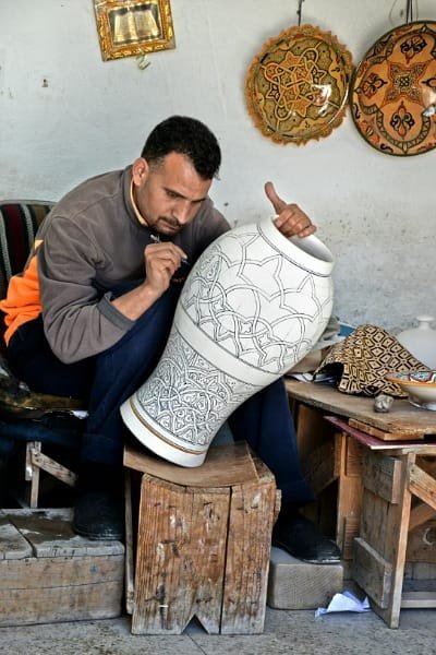 A Moroccan craftsman meticulously hand-painting intricate geometric designs onto a large ceramic vase in a workshop in Fez. The image highlights the brilliance and creativity of Moroccan artisans in pottery and traditional decorative arts.