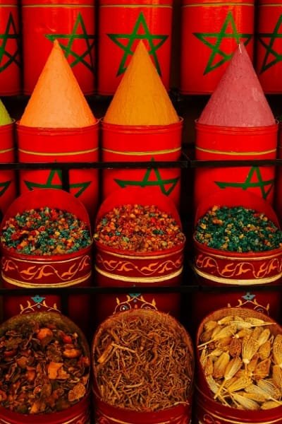 A colorful display of Moroccan spices in a Fez souk, featuring vivid red containers filled with Ras el Hanout, saffron, and other aromatic herbs. The spices are shaped into traditional cones, representing essential ingredients for Moroccan cooking and popular foodie souvenirs.