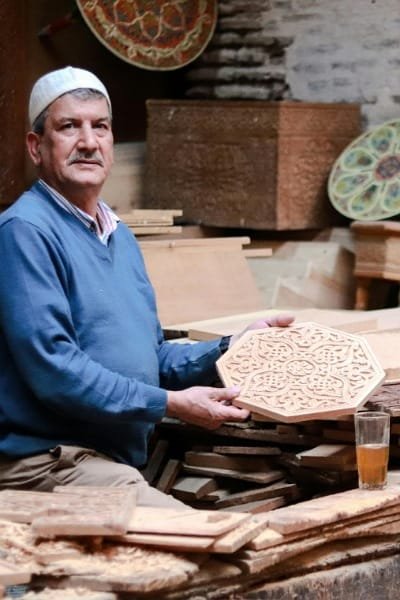 A Moroccan wood artisan holding a finely hand-carved piece of cedarwood in his workshop in Fez. The piece features intricate geometric patterns, showcasing the traditional woodcraft specialty of aromatic cedar and thuya.