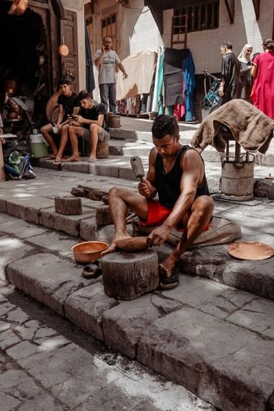 A coppersmith artisan (Saffarine) seated in the historic Place Seffarine in Fez, hand-hammering a copper piece. The scene captures the rhythmic sound and living heritage of the Coppersmiths' Souk, a traditional craft near the Al-Qarawiyyin Library.