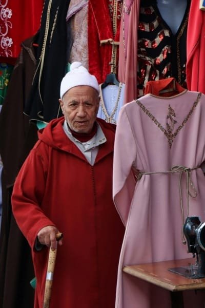 A local merchant in Fez stands beside a display featuring a traditional Moroccan caftan. The caftan is adorned with Fez embroidery, a traditional craft known for its elegance and intricate silk stitching and high craftsmanship.