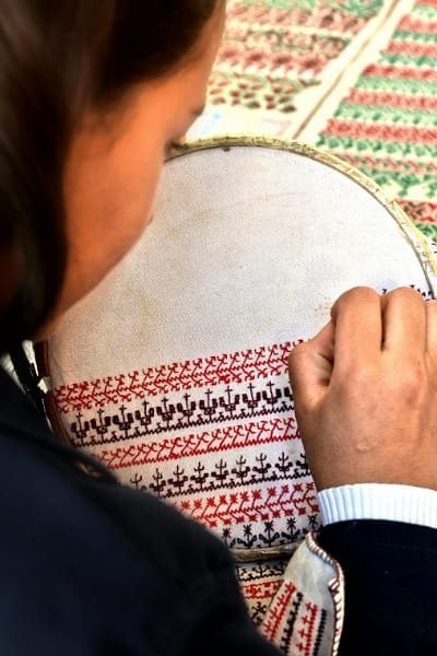 Close-up of a Moroccan artisan's hands meticulously working on a piece of Fez embroidery, using the distinctive single-faced technique. The silk threads form intricate geometric patterns, reflecting the elegance and patience required for this traditional craft.