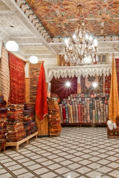 A richly decorated interior of a rug shop in Fez, displaying stacks of handwoven Moroccan carpets and rugs. The image highlights the colorful designs and traditional luxury of Berber rugs and urban-style carpets.